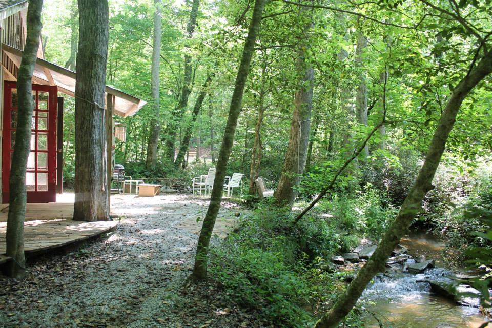 A serene forest scene featuring a cabin with red French doors, surrounded by lush green trees. A winding gravel path leads to a small stream with gently flowing water, complemented by a few white outdoor chairs positioned nearby for relaxing in nature. Sunlight filters through the foliage, creating a peaceful atmosphere. Hilltop Bike Park mountain bike trail.