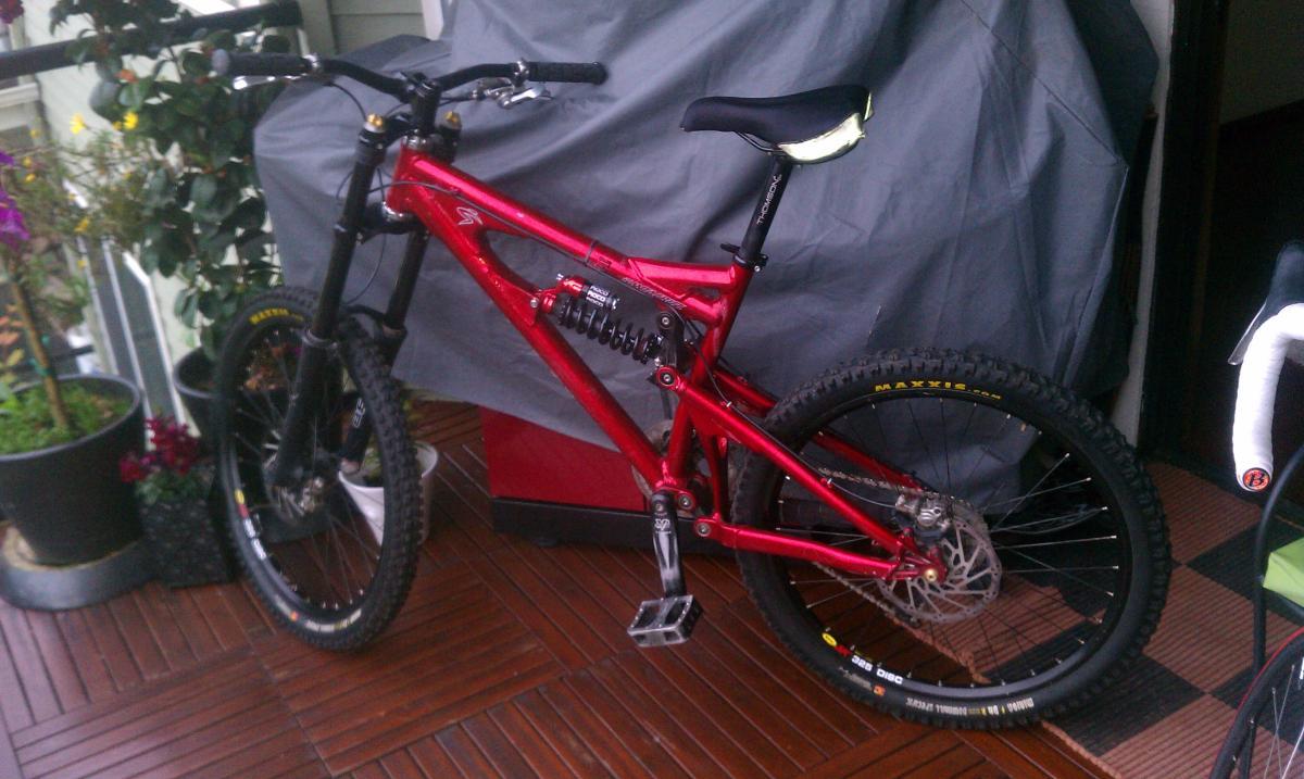 Santa Cruz Nickel: A red mountain bike with a dual suspension system is parked on a wooden deck, surrounded by potted plants. A gray tarp partially covers an object in the background. The bike features thick, knobby tires and a black seat, ready for outdoor cycling.