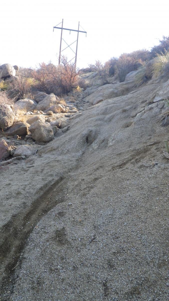 A sloped, rocky trail covered in loose sand and gravel, leading up to a power line tower in the background. Sparse vegetation and large boulders are visible along the sides of the path under a bright sky. South Foothills mountain bike trail.