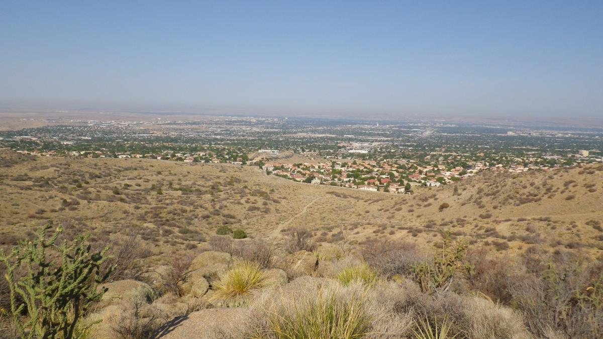 A panoramic view of a valley from a higher elevation, showcasing a mix of urban development and natural landscape. The foreground features dry, sparse vegetation and rocky terrain, while the background reveals a sprawling city with numerous houses and a clear blue sky above. South Foothills mountain bike trail.