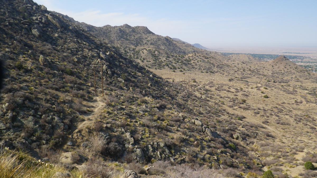 A panoramic view of rugged hills and mountains, featuring sparse vegetation and rocky terrain under a clear blue sky. Power lines are visible along a dirt path that winds through the landscape, which stretches into the distance. South Foothills mountain bike trail.