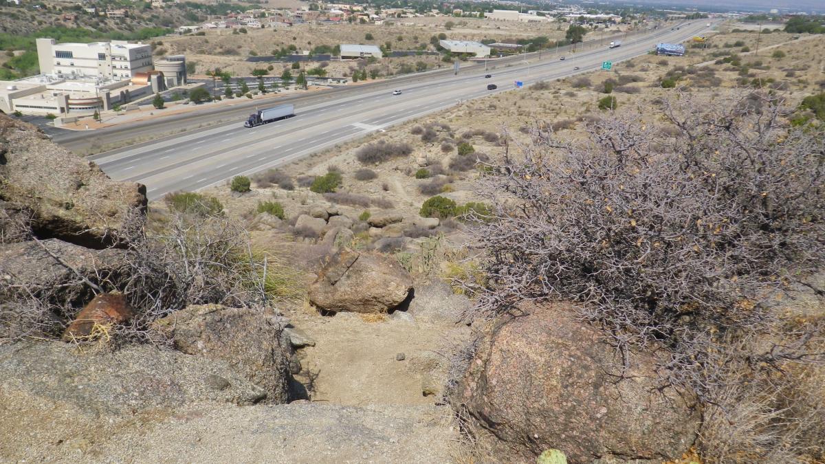 A rocky hillside with dry vegetation overlooking a highway. A truck is driving along the road, with buildings and trees visible in the background. The scene conveys a blend of natural and urban landscapes under a clear blue sky. South Foothills mountain bike trail.