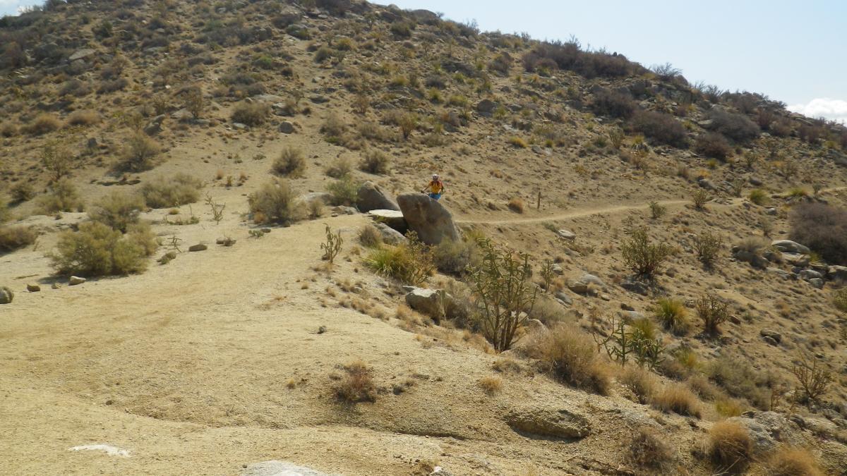 A rocky hillside with sparse vegetation, featuring a dirt path winding through the landscape. A person in a colorful shirt is sitting on a large rock near the path, surrounded by dry, desert-like terrain under a clear blue sky. South Foothills mountain bike trail.