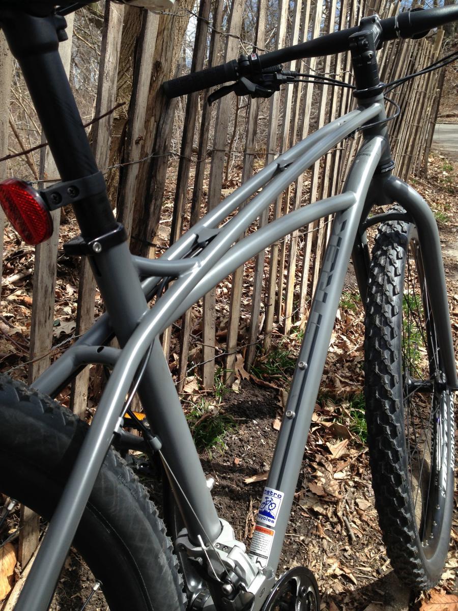 Trek Sawyer: A close-up view of a gray mountain bike resting against a wooden fence, showcasing its frame, tires, and rear wheel. The ground is covered with fallen leaves, and patches of grass are visible nearby.