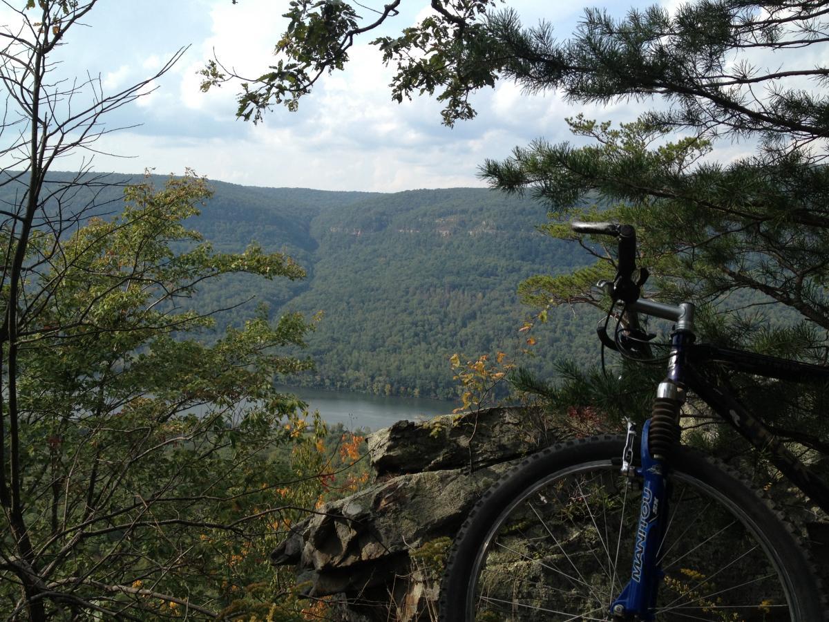 A mountain bike resting on a rocky outcrop with a scenic view of rolling green hills and a river below, framed by trees and a partly cloudy sky. Raccoon Mountain Trail Network mountain bike trail.