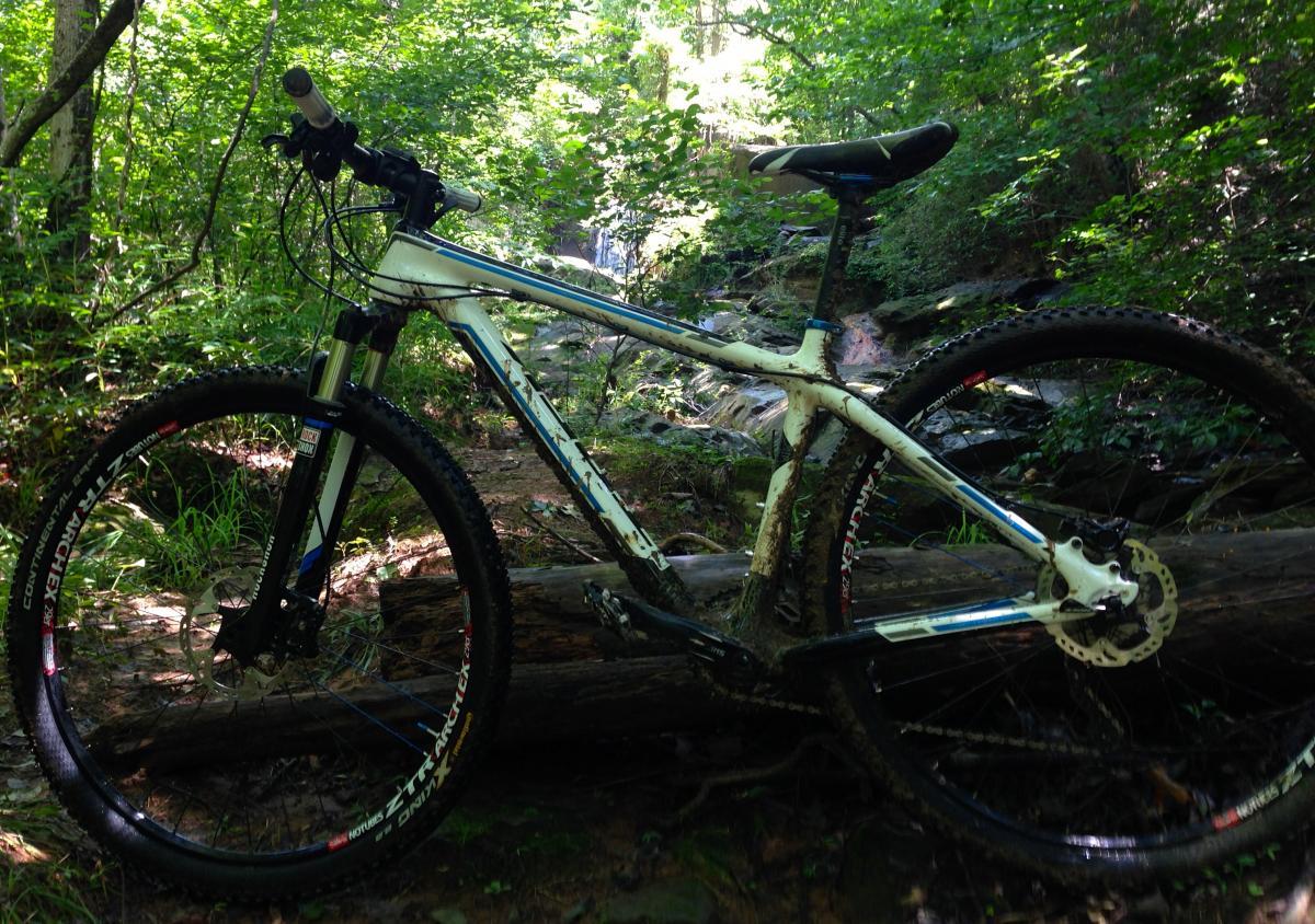 Trek Superfly Full Suspension: A mountain bike resting on a log in a lush, green forest with a small waterfall in the background, showcasing muddy tires and a well-used appearance.