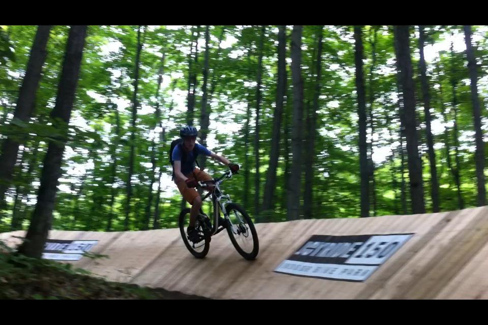 A mountain biker riding on a wooden ramp through a forested area, featuring lush green trees in the background. The cyclist is wearing a helmet and a blue shirt, with a backpack, and appears to be mid-jump or descending the ramp. A sign on the side of the ramp is partially visible. Hardwood Ski and Bike mountain bike trail.