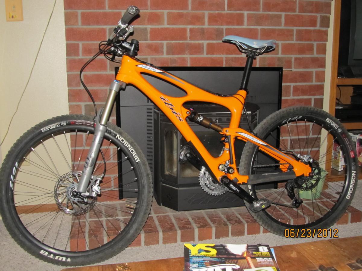 Ibis Mojo SL: A bright orange mountain bike leaning against a brick wall with a fireplace. The bike features a front suspension fork, thick tires, and disc brakes. A magazine lies on the floor in front of the bike, and there is furniture visible in the background.