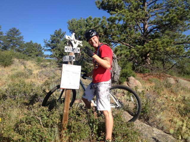 A person wearing a helmet and a red shirt stands beside a mountain bike, giving a thumbs-up gesture. They are positioned next to a trail sign that reads "Skin & Bines" in a natural outdoor setting with trees and scrub vegetation. The scene is sunny and depicts a recreational biking environment. Curt Gowdy State Park mountain bike trail.