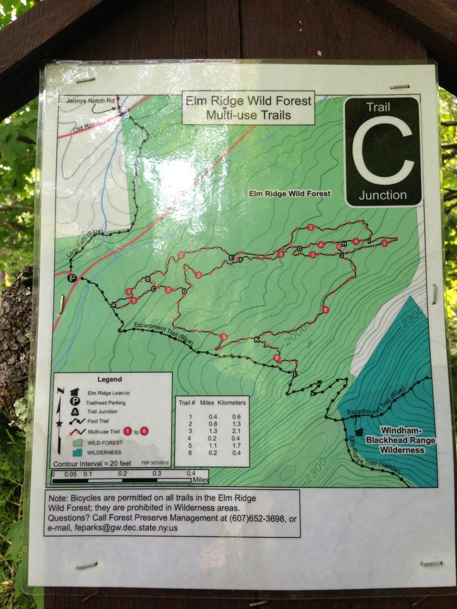A map of the Elm Ridge Wild Forest, displaying multi-use trails, including trail junctions, parking areas, and a legend for orientation. The map features topographical lines indicating elevation changes and lists the distances of various trails in both miles and kilometers. A note at the bottom mentions regulations regarding bicycle use on the trails. Elm Ridge mountain bike trail.