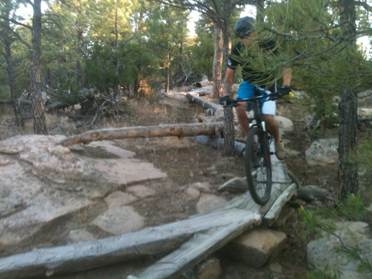 A mountain biker navigating a narrow wooden bridge over rocky terrain in a forested area, surrounded by trees and natural foliage. Glendo State Park mountain bike trail.