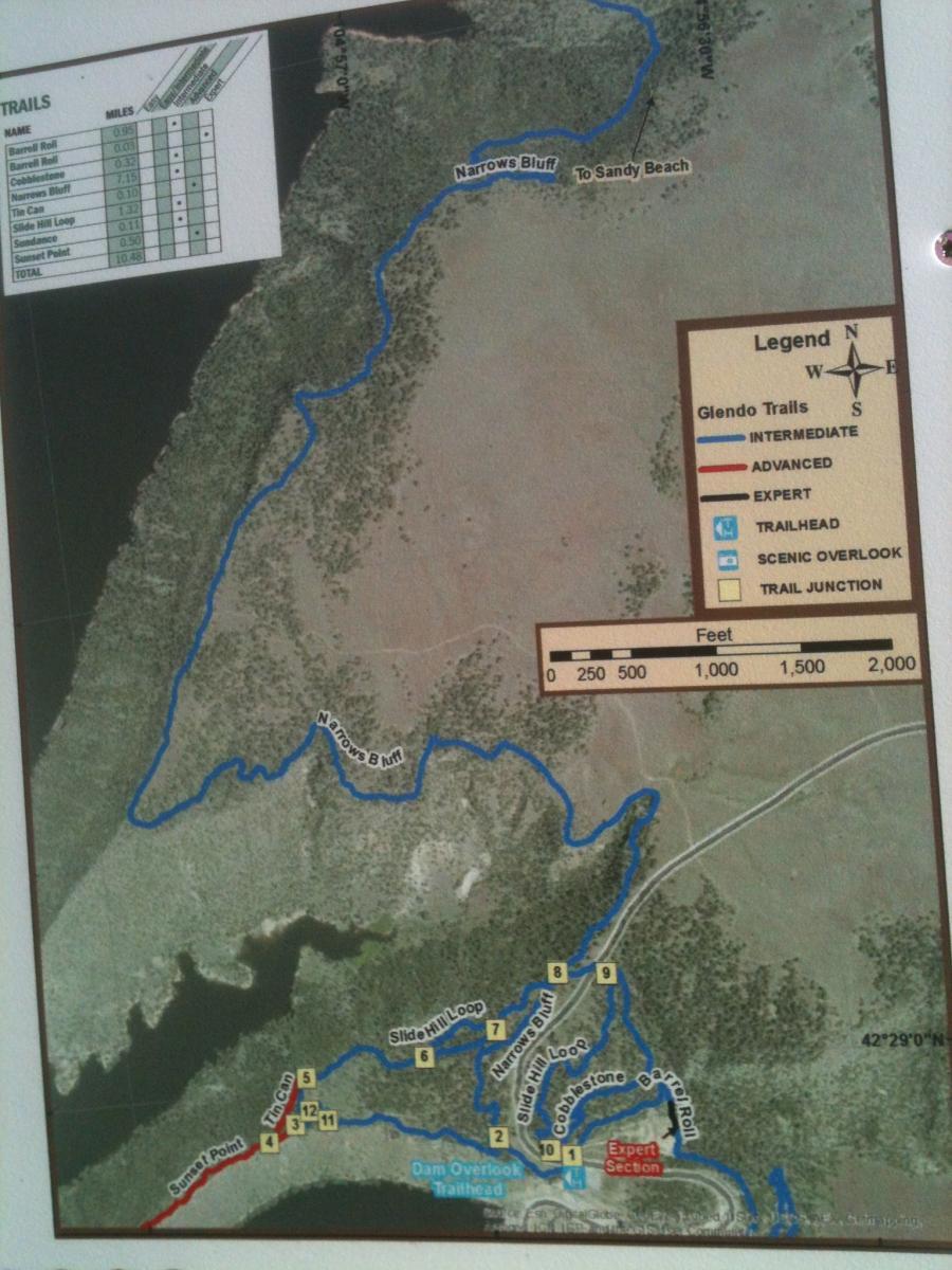 A detailed map of Glendo Trails, featuring various hiking and biking routes marked in blue. The map indicates trail names, difficulty levels (intermediate, advanced, expert), trail junctions, scenic overlooks, and trailheads. Key locations such as Narrows Bluff and Sunset Point are labeled, with additional geographical features like a lake. A legend and directional indicators are included to assist users in navigating the trails. Glendo State Park mountain bike trail.
