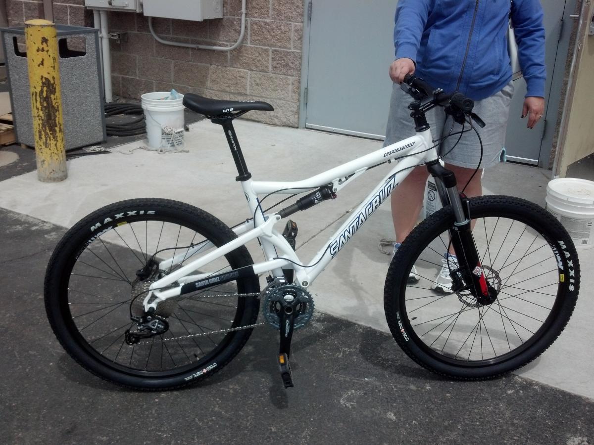 Santa Cruz Superlight: A person standing next to a white mountain bike with black wheels and the "Santa Cruz" logo visible. The bike features a dual suspension design and is positioned on a concrete surface with various background elements, including a yellow post and a bucket. The person is wearing a blue hoodie and gray shorts.
