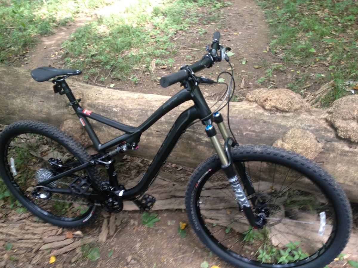 Specialized Stumpjumper FSR Comp 29: A black mountain bike resting against a fallen log in a wooded area. The bike features a suspension fork, knobby tires, and a sleek frame, surrounded by green grass and scattered leaves on the ground. A dirt trail is visible in the background.