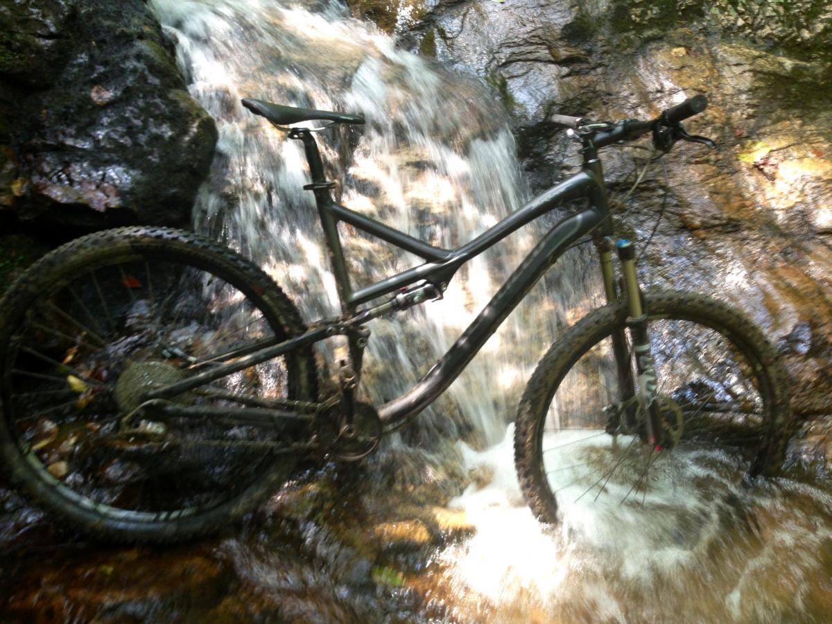 Specialized Stumpjumper FSR Comp 29: A mountain bike partially submerged in a flowing stream, with a small waterfall in the background and rocks surrounding the water. The bike appears muddy, suggesting recent use on rugged terrain. Sunlight is shining through the trees, creating a natural, outdoor setting.
