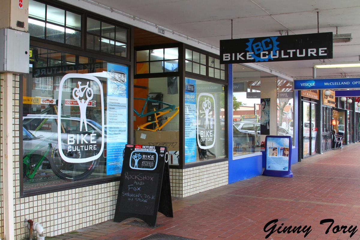 A storefront of "Bike Culture," featuring a large logo with a bicycle graphic in the window. The shop displays posters with various bike services offered, including a sign on an A-frame board that promotes servicing for RockShox and Fox products. The exterior has a tiled base and is situated along a street with other shops visible in the background.