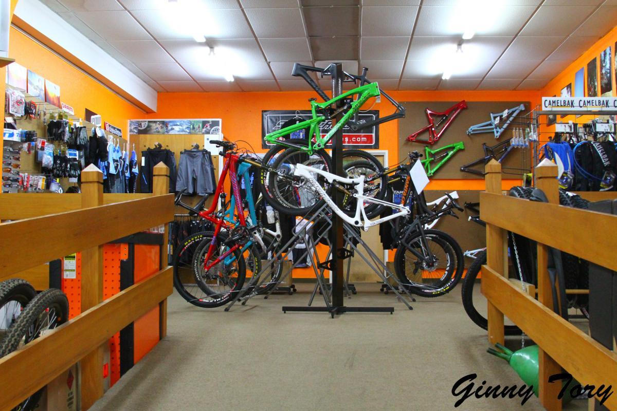 A brightly lit bicycle shop interior featuring a variety of mountain bike frames and accessories displayed on racks. The walls are painted orange and adorned with cycling gear, including clothing and equipment. A wooden railing runs along the foreground, adding to the shop's welcoming atmosphere.