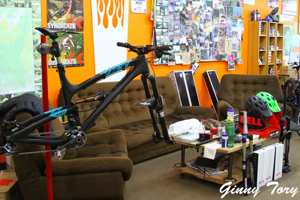A workspace featuring a bike frame on a repair stand, surrounded by various biking gear and tools. The background includes an orange wall decorated with biking posters and a collage of photos. Two brown sofas are positioned nearby, with a coffee table cluttered with drinks and supplies.