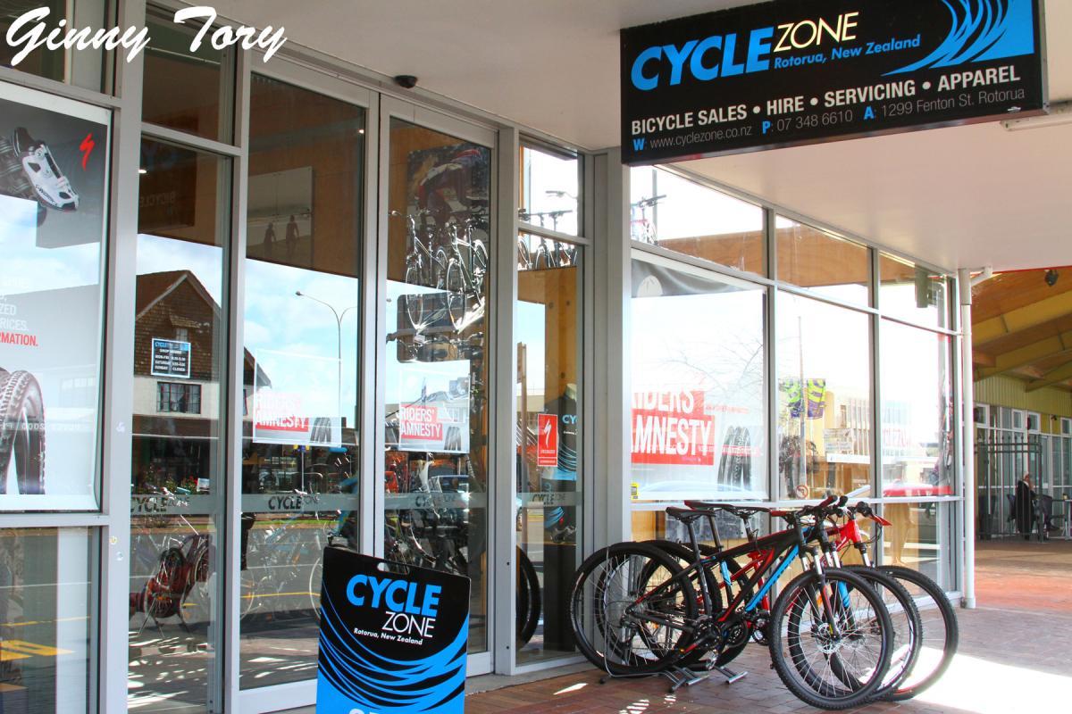 A storefront of Cycle Zone in Rotorua, New Zealand, featuring large glass windows displaying bicycles and related items. The shop has a prominent sign indicating bicycle sales, hire, servicing, and apparel. Several bicycles are parked in front, and promotional materials can be seen in the windows.