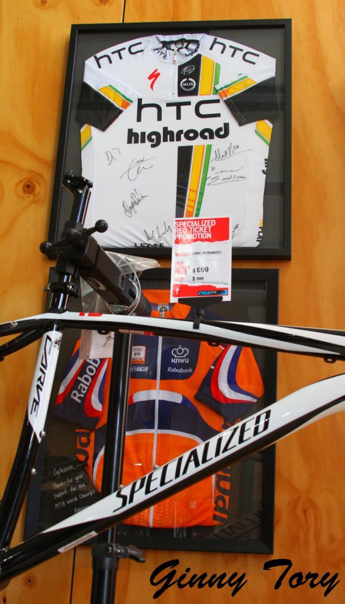 A framed cycling jersey from HTC Highroad, signed by team members, is displayed on the wall alongside a vibrant orange and blue Rabobank cycling jersey. In the foreground, a black and white Specialized bicycle frame rests on a stand, with a promotional price tag visible. The background features a wooden panel.