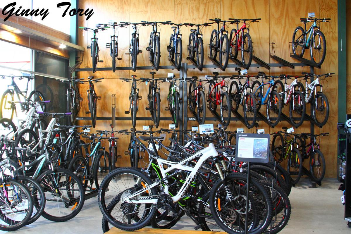 A bicycle shop interior showcasing a variety of mountain bikes. The wall features several rows of bikes displayed on racks, while the foreground presents additional bikes on display. Pricing and information tags are visible near some of the bikes, with a wood panel background and natural lighting illuminating the space.