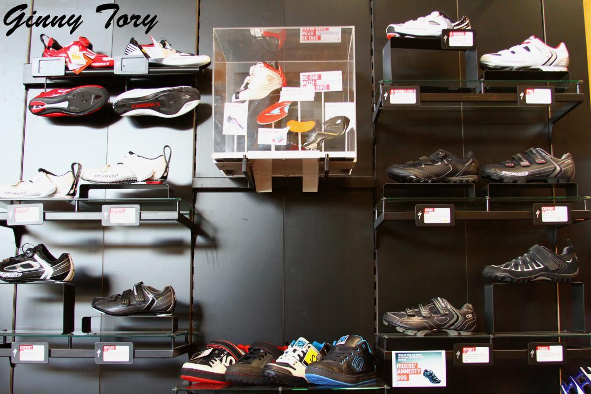 A display wall featuring various styles of athletic shoes showcased on glass shelves. The shoes include a mix of colors and designs, with several pairs prominently labeled. A transparent display case in the center holds additional footwear. The background is a dark color, highlighting the shoes.