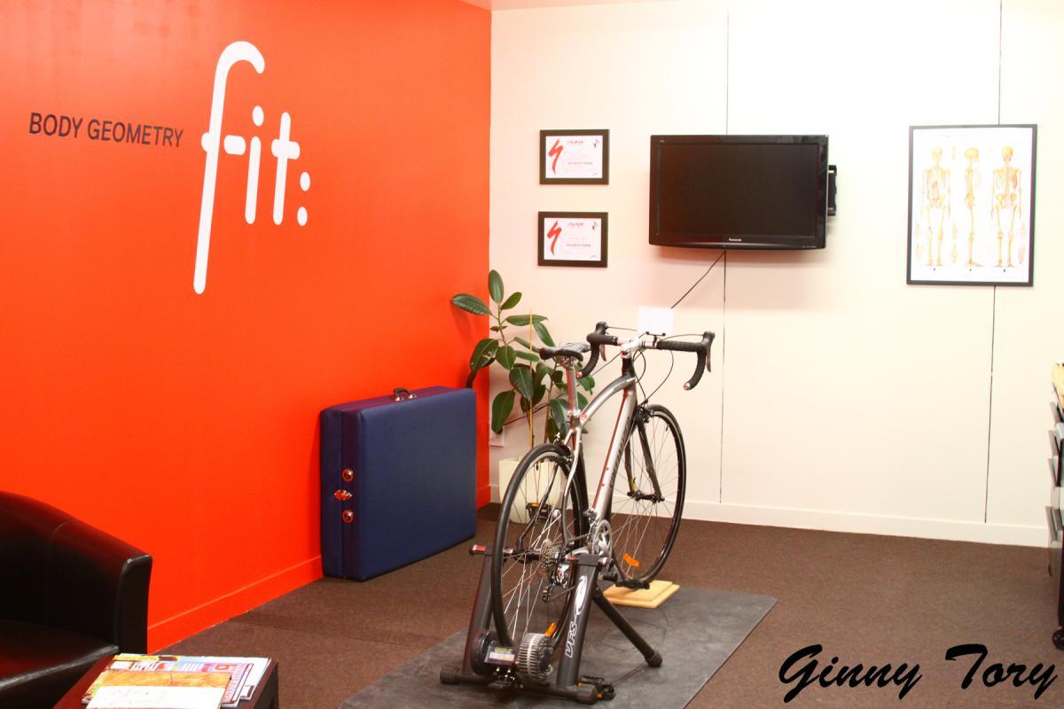 A modern fitness consultation room featuring a bright red wall with the text "BODY GEOMETRY fit:". The room includes a road bicycle on a trainer, a potted plant, a blue case, a couch, and a television mounted on the wall. Framed certificates and an anatomical poster are also visible in the background.