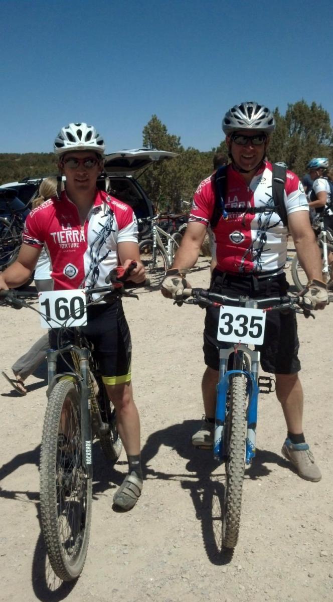 Trek Fuel EX 8: Two men stand next to their mountain bikes, both wearing matching red and white cycling jerseys with "La Tierra Torture" printed on them. They are positioned in a dirt area, likely at a biking event, with several other bicycles visible in the background. Each cyclist has a number pinned to their jerseys, with the numbers 160 and 335. The scene is set under a clear blue sky.
