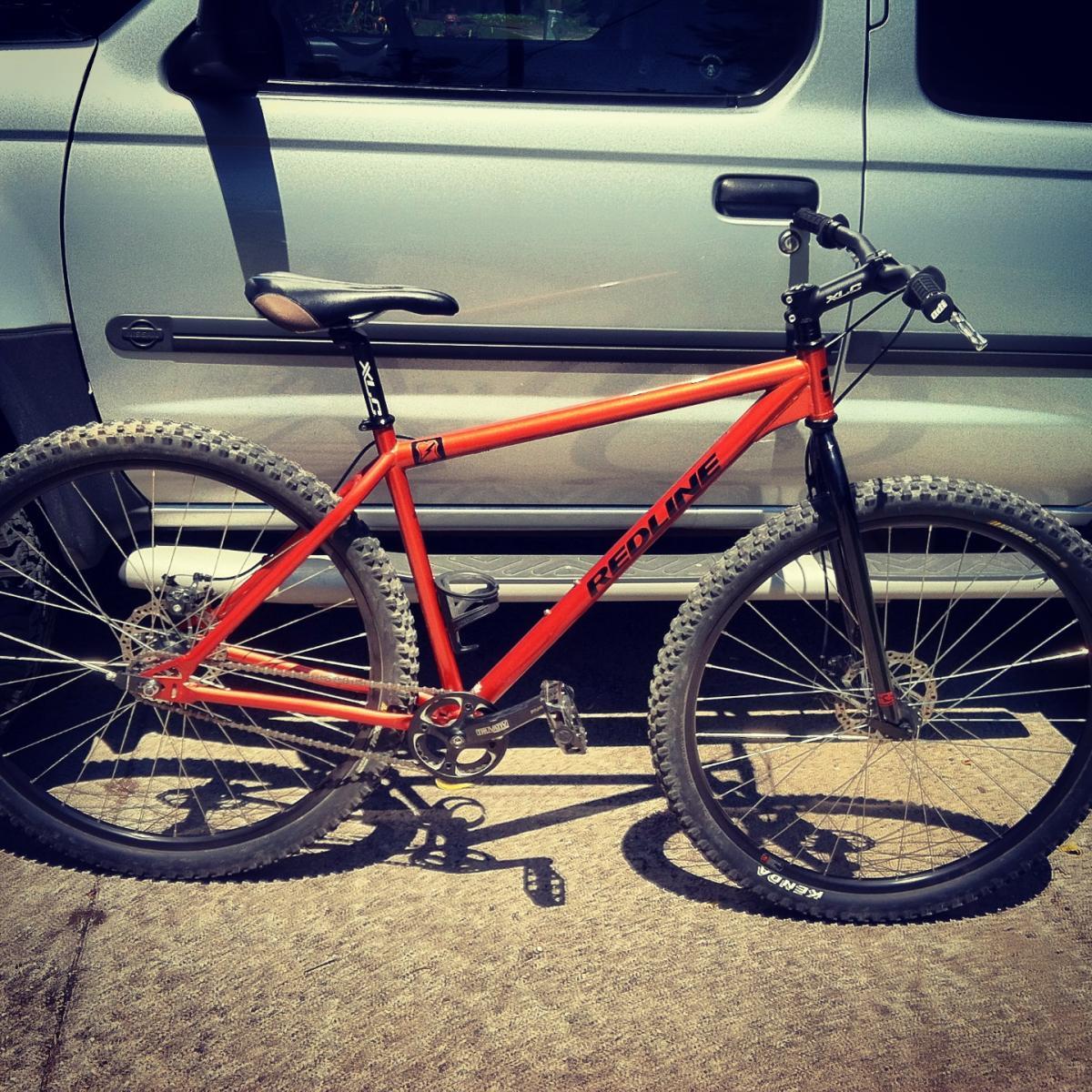 An orange Redline mountain bike parked next to a silver vehicle, featuring thick tires and a traditional frame design, resting on a textured surface.