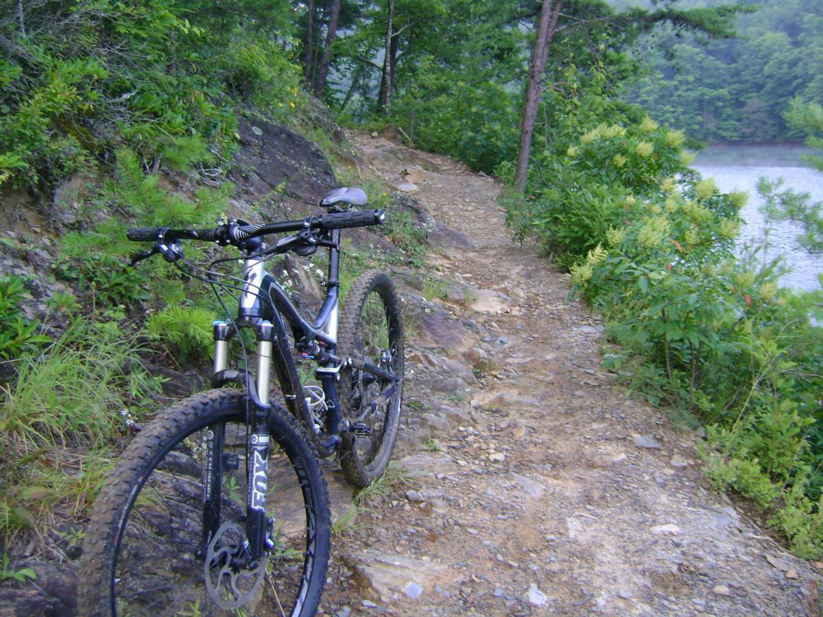 A mountain bike resting on a rocky trail surrounded by lush greenery, with a serene lake visible in the background. Tsali Left Loop mountain bike trail.