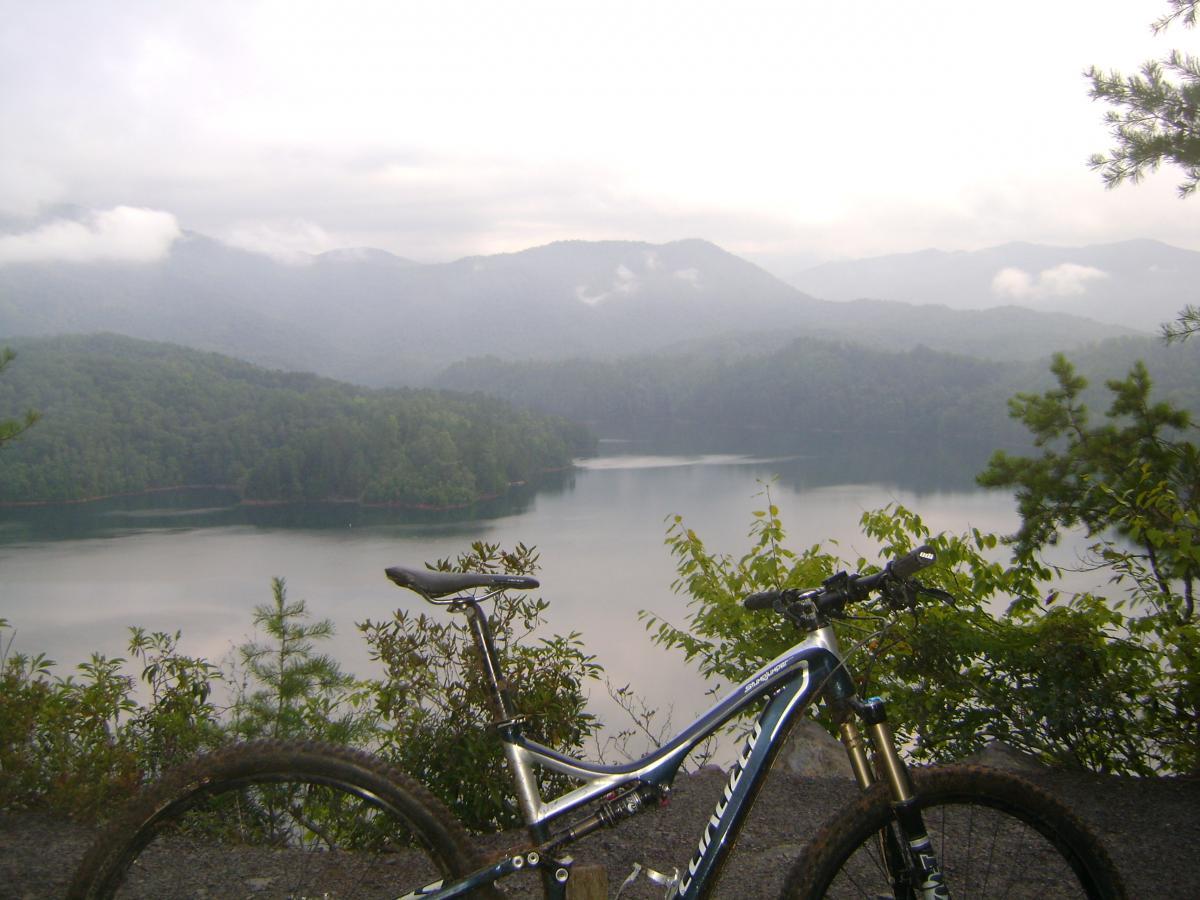 A mountain bike rests on the edge of a scenic overlook, with a serene lake and forested mountains in the background. The scene is shrouded in mist, creating a tranquil and atmospheric landscape. Tsali Left Loop mountain bike trail.