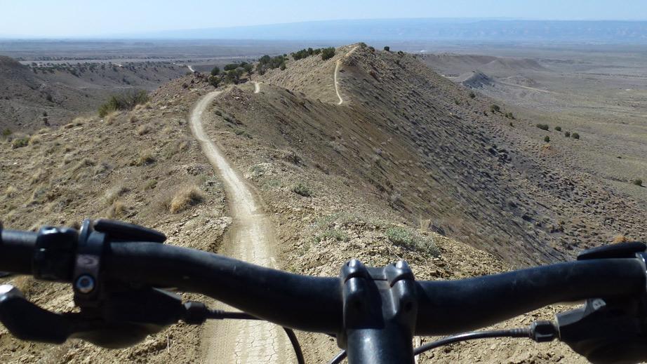 Santa Cruz Superlight: A view from the handlebars of a mountain bike, displaying a winding dirt trail along a ridge with arid landscape and distant mountains in the background.