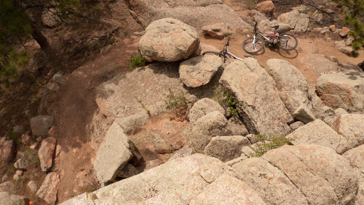 Aerial view of a rocky mountain biking trail with two bicycles resting on large boulders, surrounded by earth and sparse vegetation. Curt Gowdy State Park mountain bike trail.