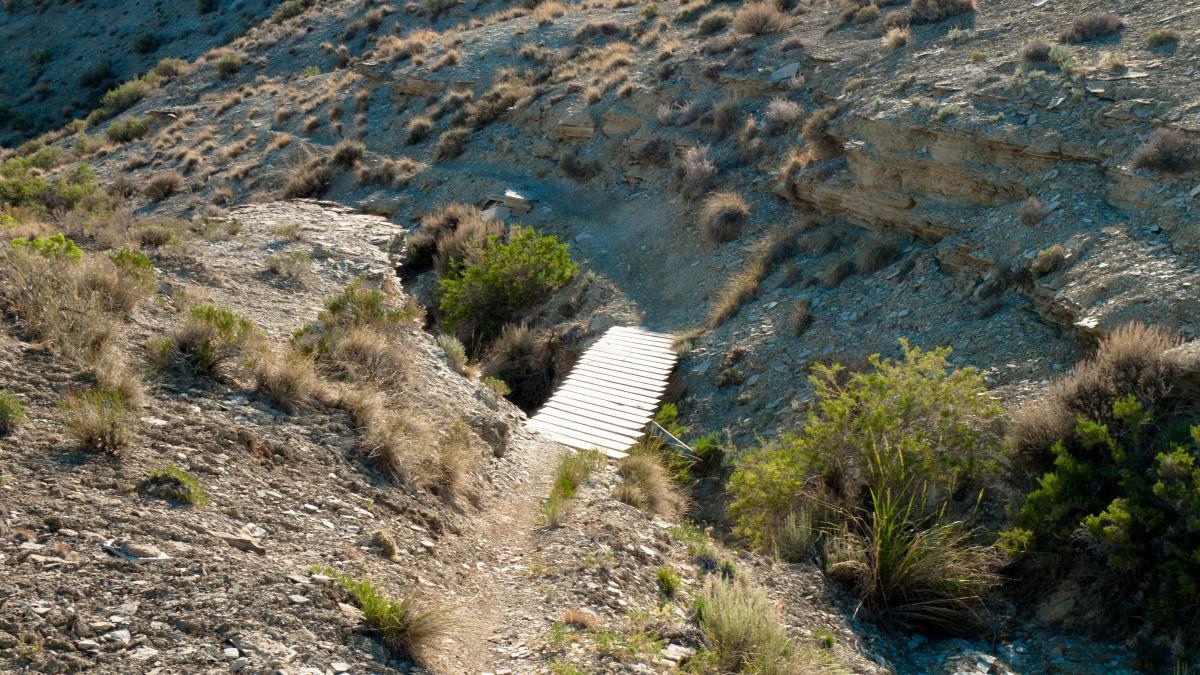 A wooden footbridge crosses a small ravine in a rocky landscape, surrounded by sparse vegetation and dry grass. The terrain is uneven, with rocky outcrops visible on both sides of the bridge. Uncle Ricos mountain bike trail.