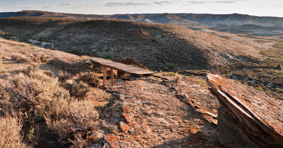 A wooden bridge extends over rocky terrain, leading into a vast landscape of rolling hills and sparse vegetation under a soft, cloudy sky. The scene captures the natural beauty of the area, with earthy tones and rugged textures highlighted by warm sunlight. Pick Your Poison mountain bike trail.