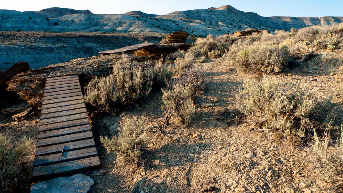 A wooden footbridge leads over rocky terrain, surrounded by shrubs and sparse vegetation, with hills in the background under a clear sky. The landscape appears dry and rugged, showcasing natural geological formations. Pick Your Poison mountain bike trail.