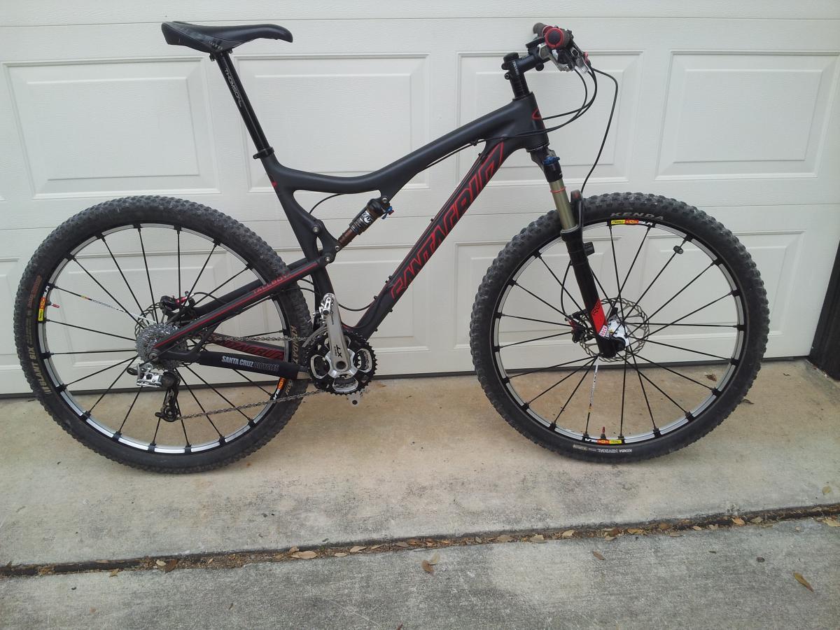 Santa Cruz Tallboy Carbon: A black and red mountain bike with mechanical disc brakes parked on a concrete surface in front of a white garage door. The bike features wide tires, a suspension fork, and a sleek frame design, showcasing its suitability for off-road trails.