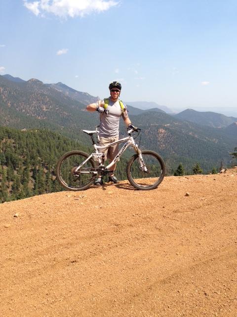 Trek Fuel EX 7: A person standing next to a mountain bike on a dirt trail, with a scenic view of mountains and a forest in the background. The individual is wearing a helmet and sporting a thumbs-up gesture, indicating a positive experience outdoors.