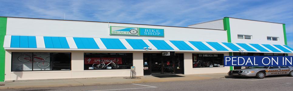A bright and inviting bike shop exterior, featuring a white façade with green accents and a blue awning. Large windows display various bicycles for sale, including a prominent red bike. A sign above the entrance reads "Beach Bike Shop." The image conveys a welcoming atmosphere for cycling enthusiasts.