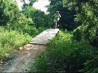 A person riding a mountain bike is mid-air over a wooden ramp on a trail surrounded by lush greenery. Loyce E. Harpe Park mountain bike trail.