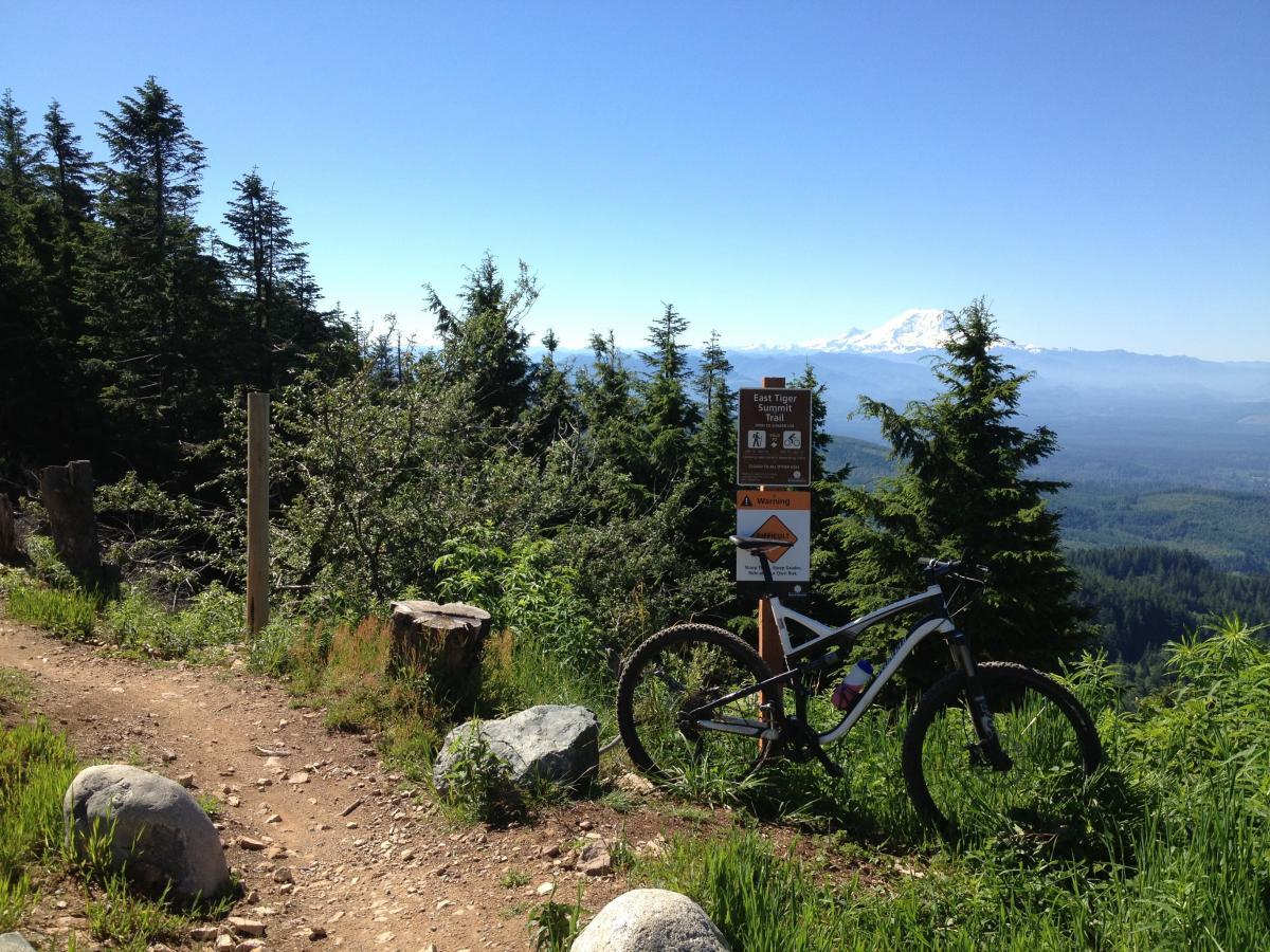 A mountain bike parked near a trail sign for the East Tiper Summit Trail, surrounded by lush green trees and a clear blue sky. In the background, there is a mountain view, adding a scenic backdrop to the biking trail. Tiger Mountain mountain bike trail.