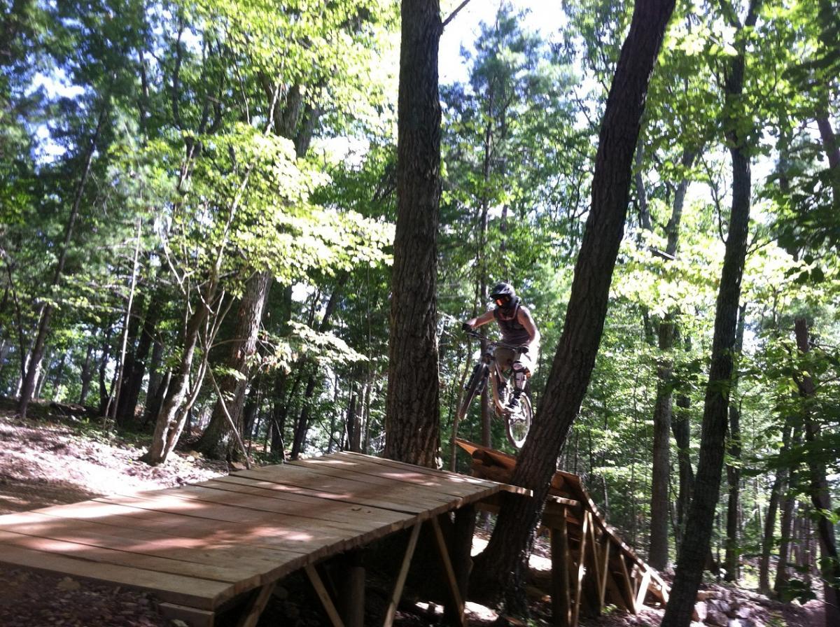 A mountain biker jumps off a wooden ramp positioned in a forested area, surrounded by tall trees and bright green foliage. The sun filters through the leaves, creating a dappled light effect on the ground. Bryce Mountain Bike Park mountain bike trail.