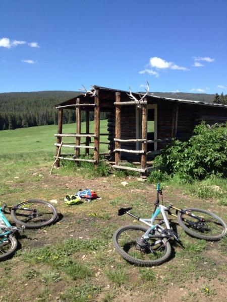 A rustic wooden cabin with antlers on the roof set in a green, open field surrounded by forested hills. Two mountain bikes lie on the ground nearby, with a helmet and some gear scattered in the grass under a clear blue sky. Meadow Mountain mountain bike trail.