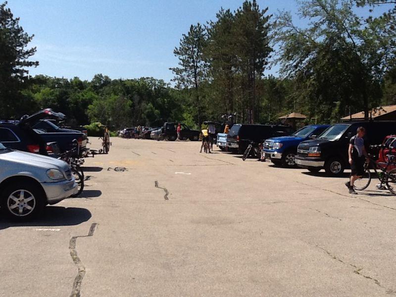 A sunny day at a parking lot filled with parked vehicles, including several SUVs and sedans, with people preparing for a biking or outdoor activity. In the background, trees provide a natural setting, and cyclists can be seen either getting ready or riding away. Kettle Moraine John Muir + Emma Carlin mountain bike trail.
