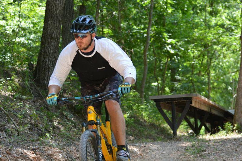 A mountain biker wearing a helmet and sunglasses rides a yellow bike along a dirt trail in a forested area, surrounded by lush green trees. A wooden bench is visible in the background. Slaughter Pen Trail mountain bike trail.