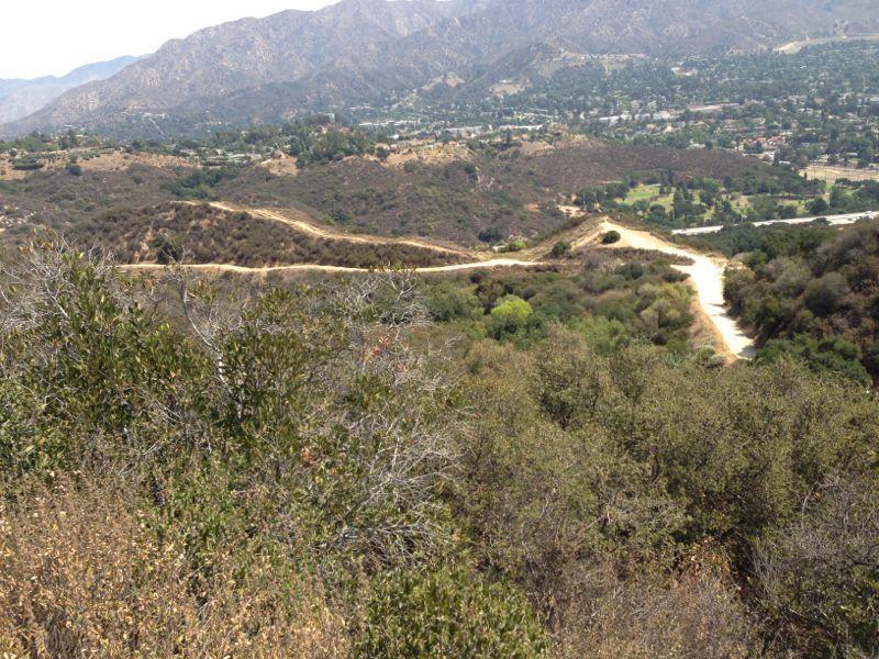 A scenic view of rolling hills and winding dirt paths, with a backdrop of mountains and a glimpse of a nearby residential area in the valley below. The landscape is mostly covered in greenery, with a mix of trees and shrubs. La Tuna Canyon mountain bike trail.