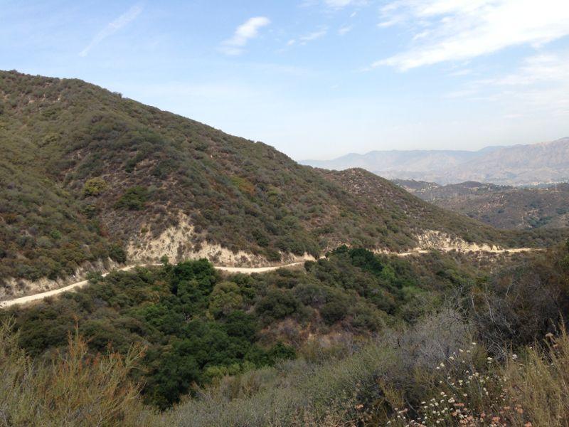 A scenic view of rolling hills covered in greenery with a dirt path winding through the landscape, set against a backdrop of distant mountains and a partly cloudy sky. La Tuna Canyon mountain bike trail.