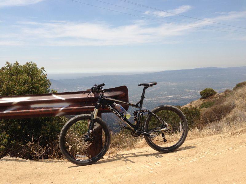 Mountain bike leaning against a guardrail, overlooking a scenic view of a valley and distant mountains under a clear blue sky. The foreground features dry grass and shrubbery, suggesting a trail or road. La Tuna Canyon mountain bike trail.