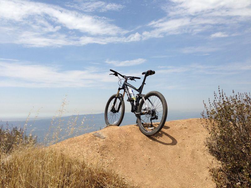 A mountain bike resting on a rocky outcrop with a clear blue sky and distant hills in the background. Dry grass and shrubs surround the bike, creating a serene outdoor setting. La Tuna Canyon mountain bike trail.