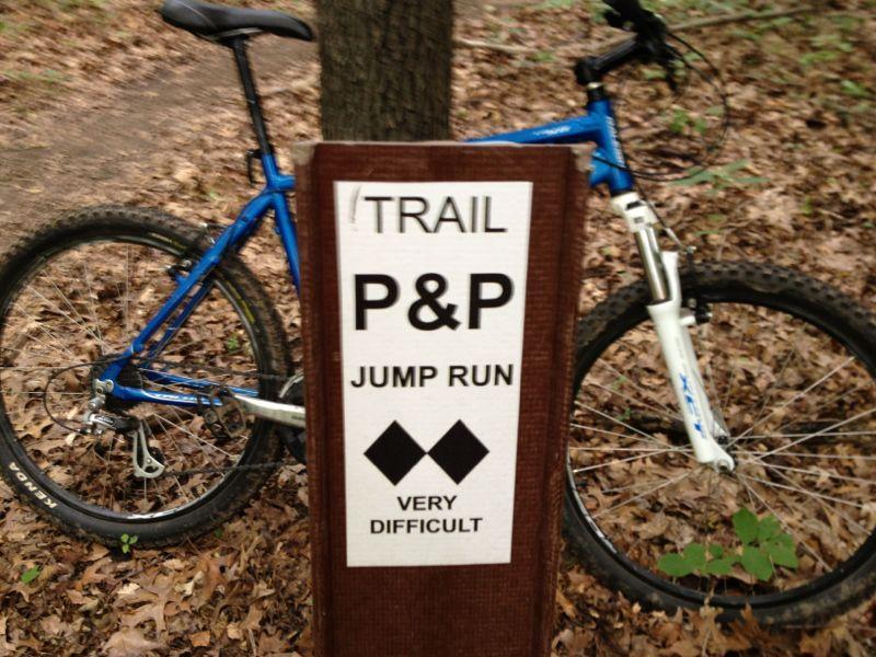 A blue mountain bike is parked next to a brown trail sign in a wooded area. The sign displays "TRAIL P&P JUMP RUN" with a symbol indicating a very difficult trail. The ground is covered with fallen leaves, and trees surround the scene. Kickapoo mountain bike trail.